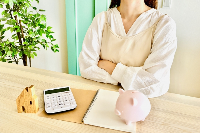 日用品の出費を抑えるために家計簿や電卓、豚の貯金箱を使って節約を考える主婦の様子。